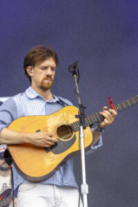 Male guitarist performing on stage, wearing a blue striped shirt and white pants, playing an acoustic guitar into a microphone on a stand.