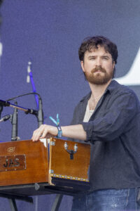 Male musician playing a wooden keyboard on stage, wearing a dark shirt and jeans, with a beard and focused expression