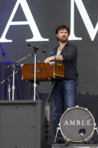 Bearded man on a stage playing a wooden keyboard-style instrument, with microphones nearby and a large backdrop behind him.