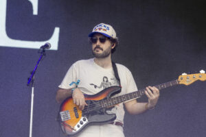 Bass guitarist performing on stage in a white t-shirt and Knicks cap, wearing sunglasses and playing a sunburst bass near a microphone stand.