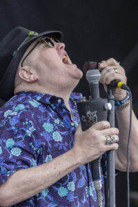 Performer in a purple floral shirt sings into a handheld microphone on stage, wearing a hat and sunglasses.