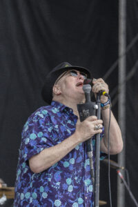 Male singer performing on stage, wearing a black hat and sunglasses, singing into a microphone while holding a phone-like device; purple floral shirt visible.