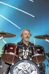 Drummer with gray hair performing behind a red drum kit on a blue stage backdrop, eyes closed and sticks in motion.