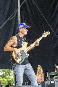 Male bass guitarist wearing a blue cap and neon sunglasses performs on an outdoor stage with a bass guitar.