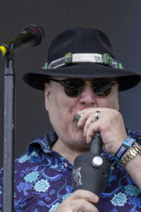 Male singer wearing a black hat and sunglasses, holding a microphone on stage with a blue floral shirt and a wristband visible