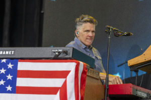 Musician playing a keyboard on stage with a patriotic flag draped over the instrument and wearing an in-ear monitor.