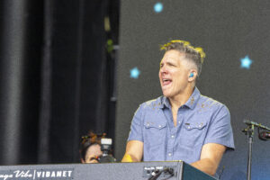 Musician on stage playing a keyboard, singing into a mic, wearing a blue shirt and blue in-ear monitor.