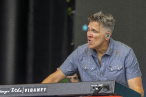 Male musician with spiky hair in a blue embroidered shirt playing a keyboard on stage, wearing a blue in-ear monitor.