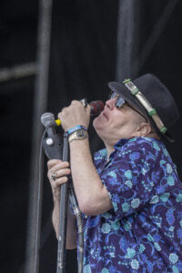 Male singer on stage wearing a black hat with green embellishments, sunglasses, and a purple floral shirt, singing into a handheld microphone.