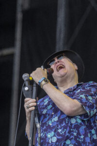 Male singer wearing sunglasses and a wide-brim hat, passionately singing into a microphone on stage.