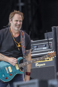 Male musician playing a turquoise electric guitar on stage, wearing beaded necklaces and a black shirt.