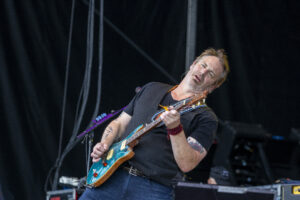 Guitarist on stage playing a turquoise electric guitar, wearing a black shirt and red beaded bracelet.