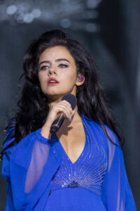 Female singer with dark hair wearing a blue outfit, holding a microphone close to her mouth on stage.
