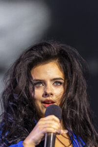 Close-up of a woman with dark hair speaking or singing into a handheld microphone.
