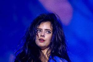 Close-up of a woman’s face with dark wet hair under blue stage lighting, looking upward.