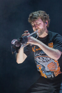 Male trumpet player in a black graphic T-shirt performing on stage, close-up with a silver trumpet.