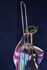 Musician raises a trombone above their head during a performance, wearing a rainbow-striped shirt against a dark background