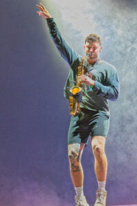 Male saxophonist performing on stage, one arm raised, saxophone held near chest with smoke-filled purple backdrop behind him.