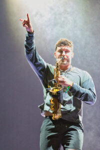 Saxophonist on stage, raising his right hand in the air while playing a brass saxophone under concert lights.