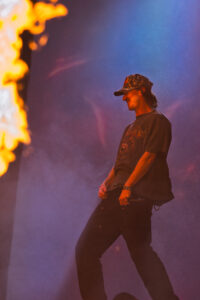 Male performer wearing a camouflage cap and dark T-shirt on stage, with a tall flame on the left and purple-blue lighting in the background.
