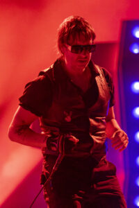Male performer on stage in red-tinted lighting, wearing sunglasses and a black vest, with a tattooed forearm visible and a microphone cable nearby.