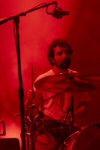 Drummer with a beard playing a drum kit on a red-lit stage, cymbals visible in front of him.