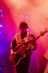 Musician playing a glittery bass guitar on a pink-lit stage, wearing glasses and a striped jacket.
