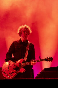 Guitarist on a red-lit stage playing a semi-hollow electric guitar, wearing a dark denim jacket and patterned strap.