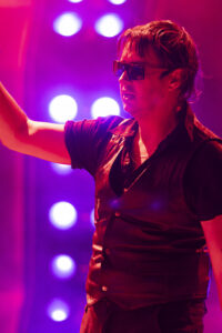 Musician wearing sunglasses on stage, raising an arm amid pink and purple stage lights.