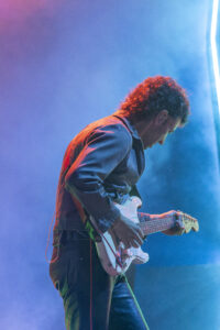 Guitarist playing a white electric guitar on a blue-tinged stage with purple lighting