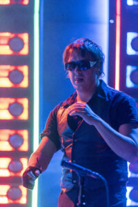 DJ wearing sunglasses performing on stage under neon blue and red lights, mid-action with hands raised