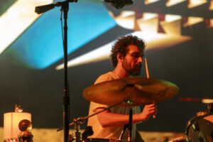 Bearded drummer in a white shirt playing drums on a brightly lit stage, holding drumsticks above a cymbal with colorful background lights.