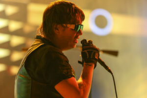 Male singer wearing reflective sunglasses, microphone in hand, performing on a warmly lit stage.