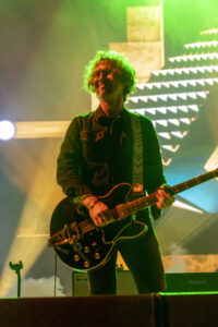 Guitarist performing on stage under green lights, playing a black electric guitar with a patterned strap.