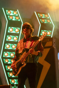 Male bassist performing on a neon-lit stage with green square light panels behind him.