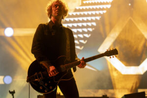 Guitarist performing on a dim stage under orange backlights, playing a black electric guitar with a striped strap.
