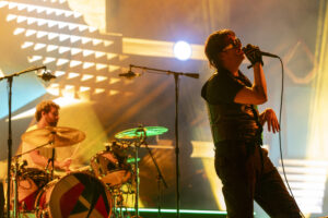 Male lead singer in black outfit and sunglasses sings into a mic on stage, with a drummer and bright stage lights behind him.