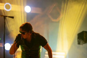 Male musician wearing sunglasses singing into a microphone on a colorful stage with warm yellow and cool teal lighting.