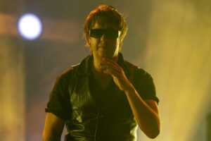 Male singer wearing dark sunglasses and a black leather vest performs on a warmly lit stage.