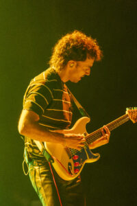 Male guitarist playing a white electric guitar on stage, lit by warm yellow lights into the audience area.