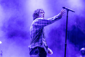 Male singer on stage reaching toward a microphone amid purple concert lighting.