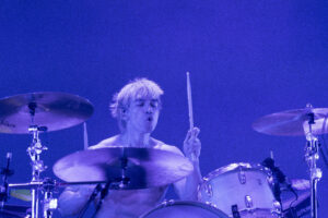 Drummer mid-performance behind a drum kit under blue stage lights, sticks raised mid-strike.
