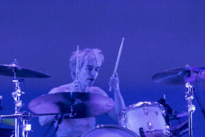 Drummer with blond hair playing a drum kit on a dim stage bathed in blue light.