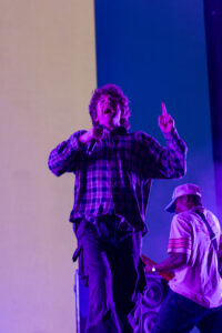 Lead singer in a plaid shirt belts into a microphone on a purple-lit stage, finger raised in the air.