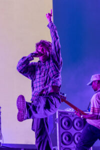 Male singer on stage, microphone in hand, one arm raised, wearing a plaid shirt under purple stage lighting.