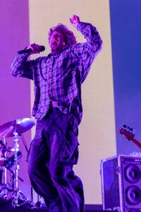 Male singer in a plaid shirt belts into a microphone on stage under purple lighting