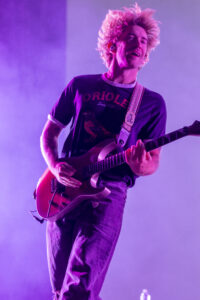 Male guitarist performing on stage, playing a red electric guitar under purple lighting, wearing a dark t-shirt and jeans.