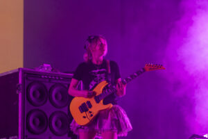 Guitarist performing on stage with bright orange guitar and purple lighting, smoke in the air, wearing a bandana and black shirt.