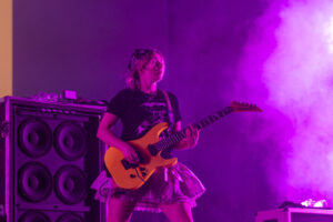 Female guitarist performing on stage with an orange electric guitar and large speaker cabinets, under purple stage lighting.