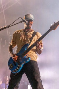 Male musician on stage playing a blue electric bass guitar, wearing a beige T-shirt and cap, microphone nearby.